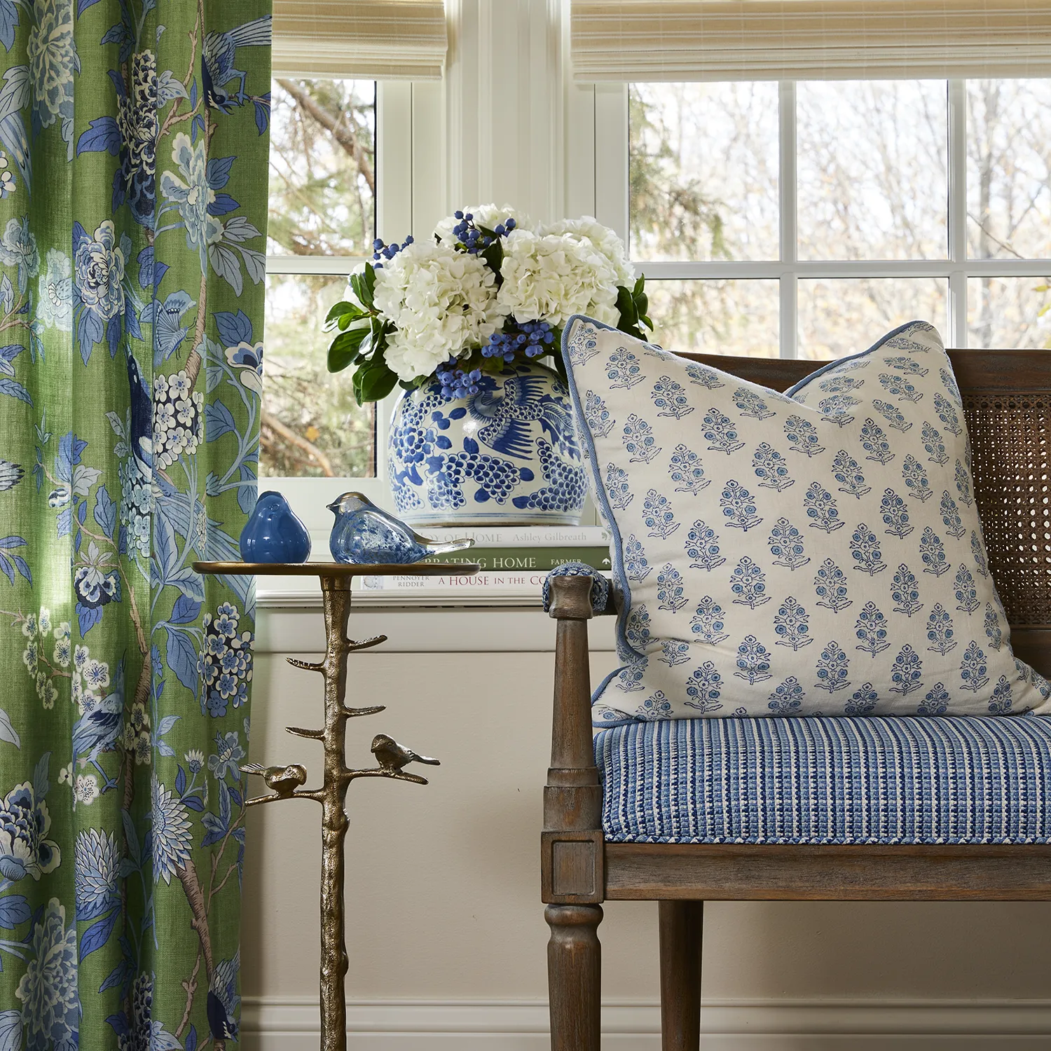 A window nook featuring green floral drapes, a patterned wooden bench with a blue throw pillow, and white hydrangeas on the windowsill.