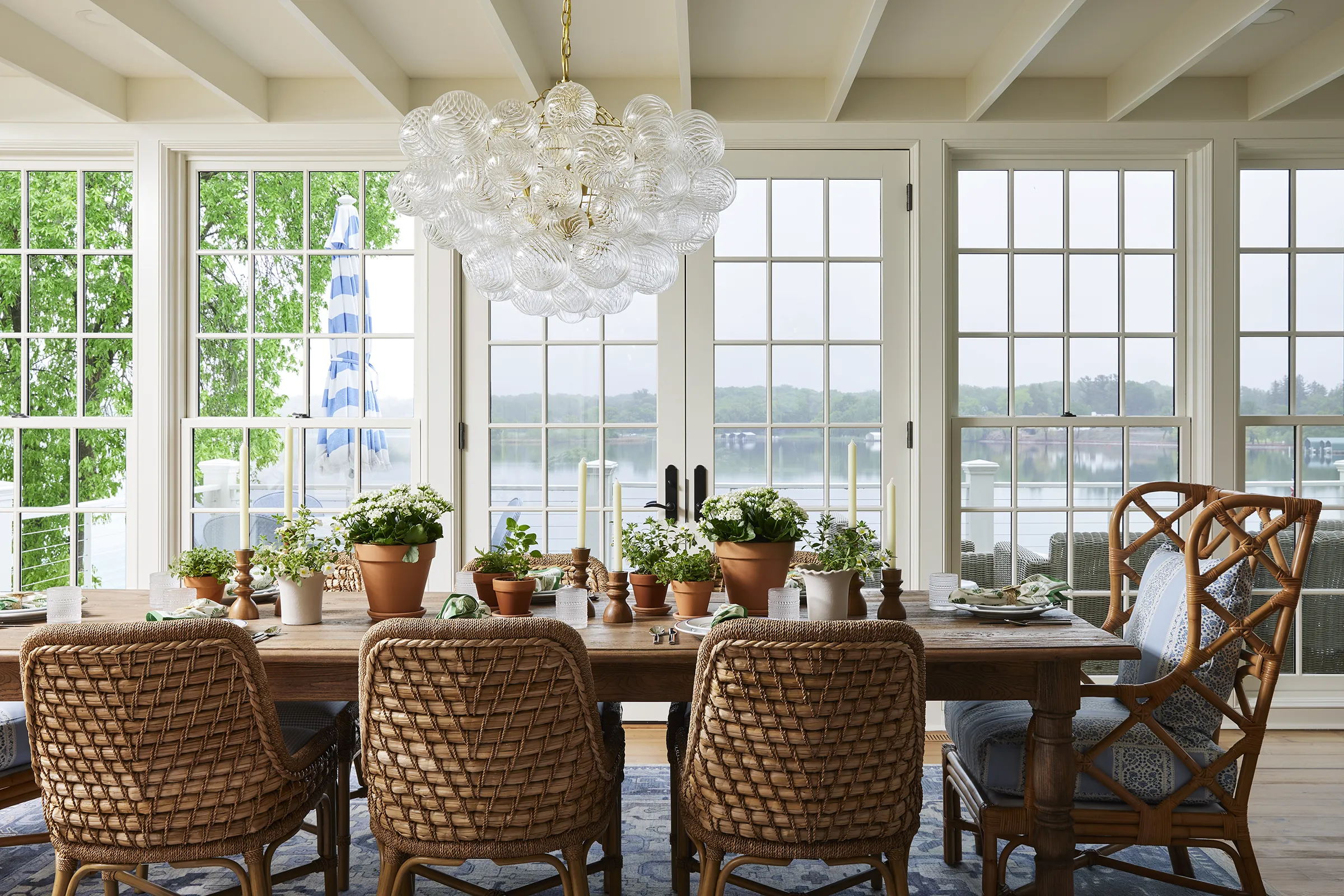 Bright sunroom dining area by Grace Hill Design with woven rattan chairs, a long wooden table set with potted plants, and a bubble glass chandelier overlooking a lake view.
