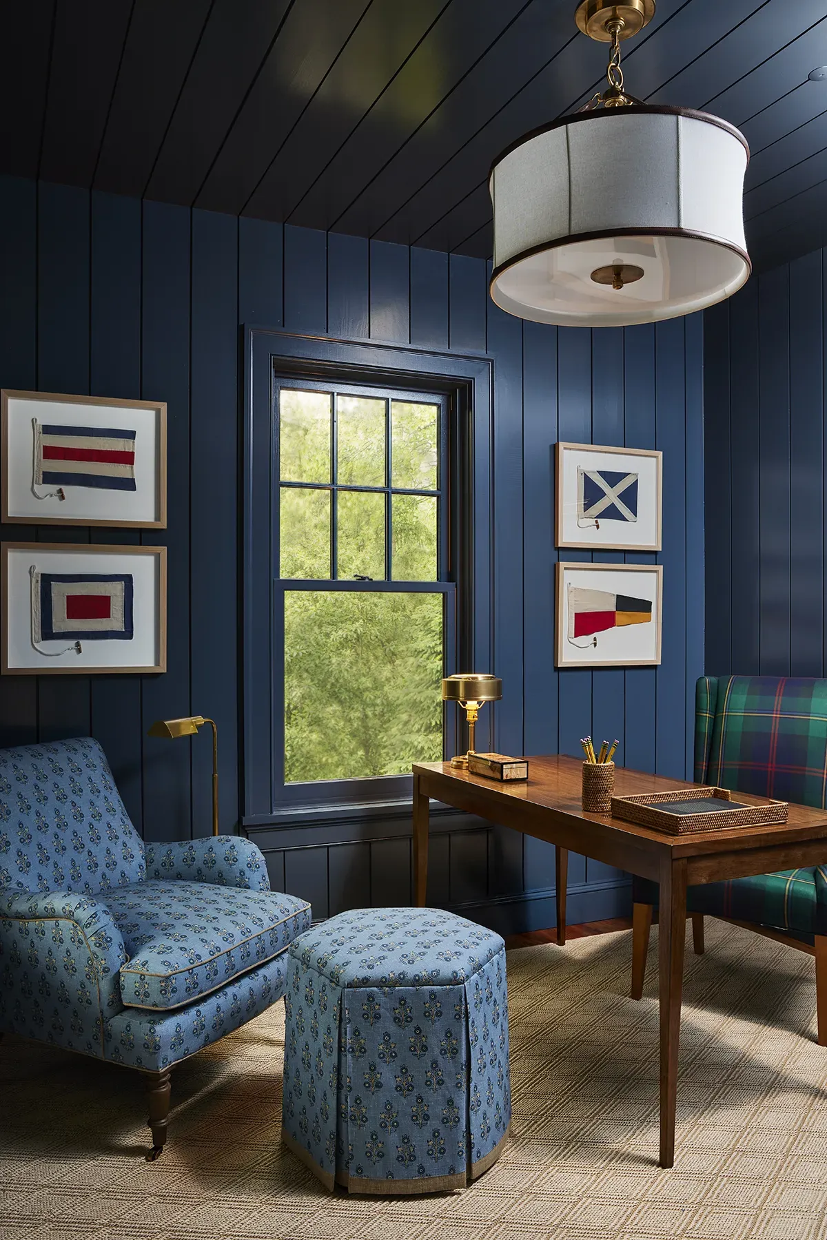 Nautical themed home office featuring dark navy blue wood-paneled walls, framed artwork, and a modern drum pendant ceiling light