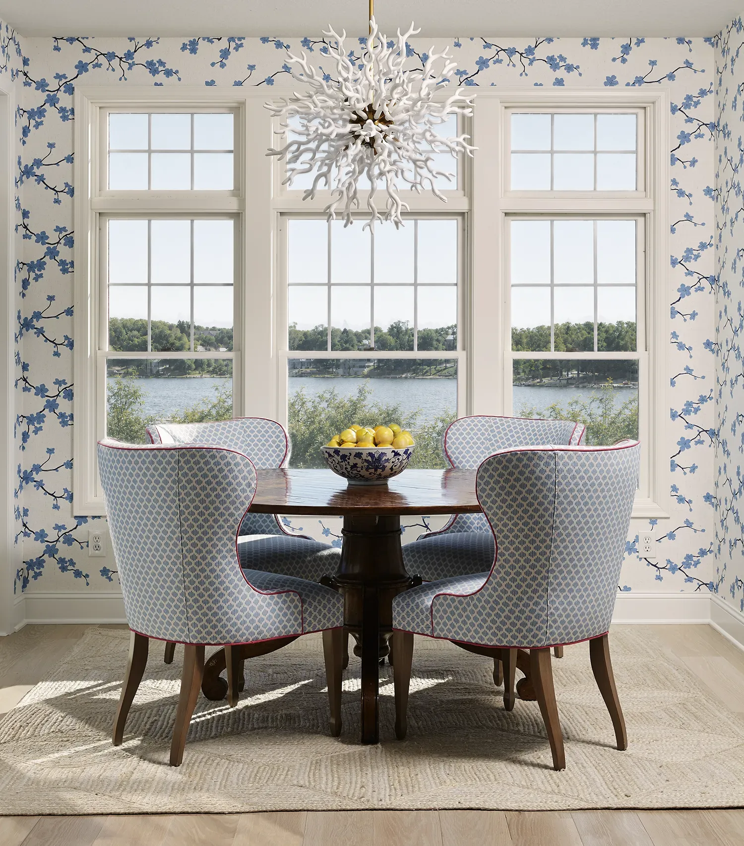 Coastal dining room with a round wooden table, blue patterned wingback chairs with coral piping, and a white coral branch chandelier