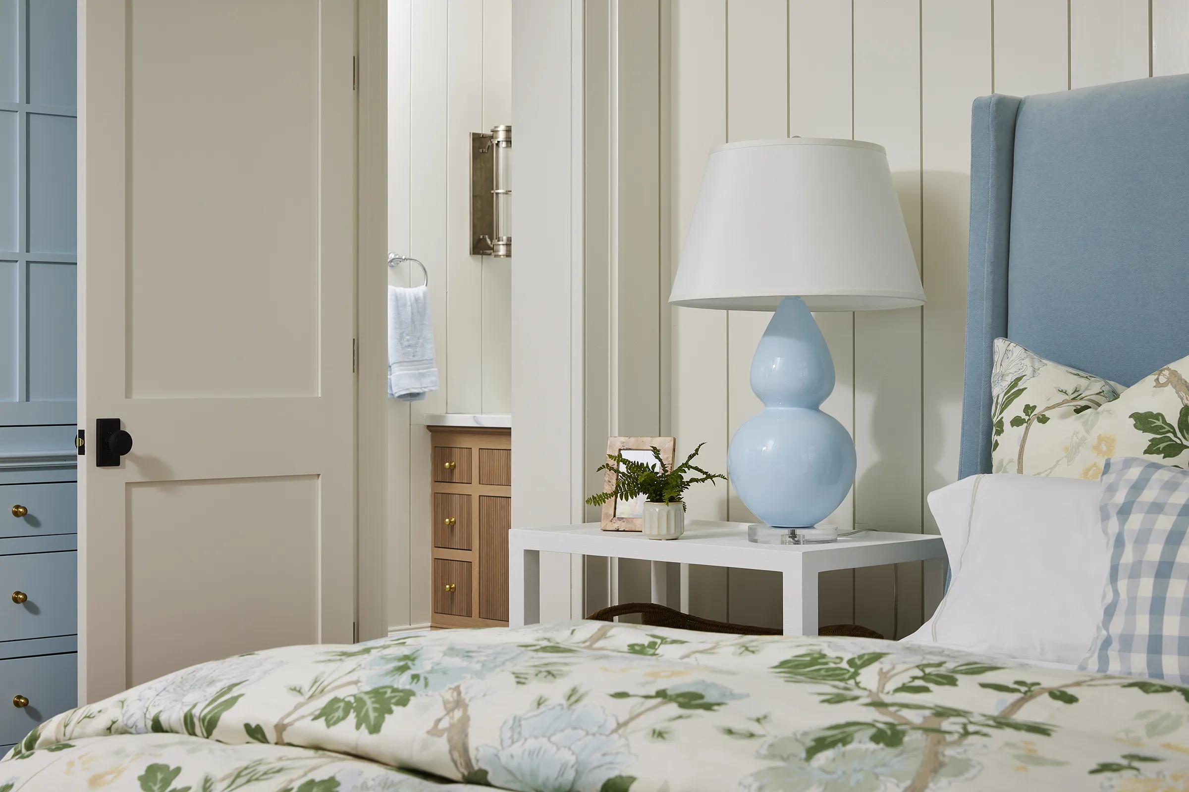 An elegant bedroom featuring a soft blue upholstered headboard, cream vertical shiplap walls, a white minimalist nightstand, and a prominent sky blue double gourd table lamp with a white tapered shade
